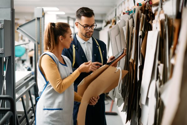 Young clothing designer and seamstress using touchpad and discussing about new clothing pattern at textile factory.
